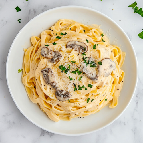 Top-down view of creamy mushroom fettuccine pasta served on a white ceramic plate over a white marble countertop. The pasta is generously coated in a rich cream sauce filled with mushrooms and parmesan, then garnished with fresh chopped parsley. The dish looks elegant, warm, and inviting, perfect for a cozy dinner.