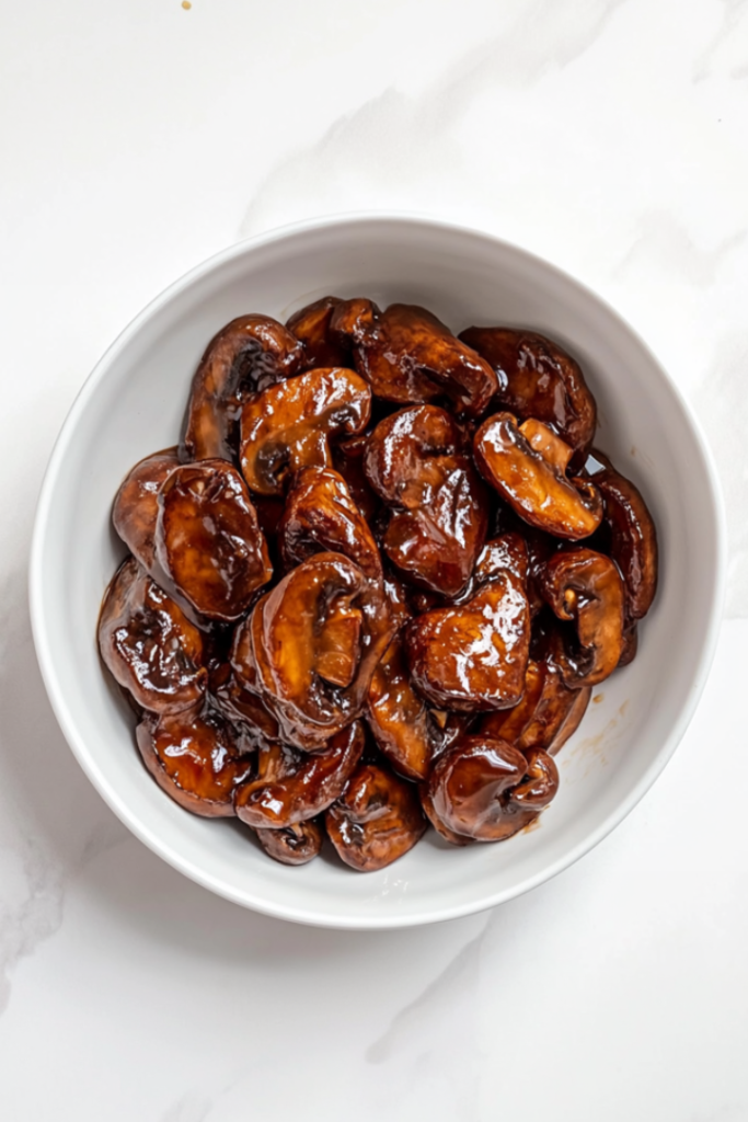 A top-down view of glossy garlic butter mushrooms served in a clean white bowl, placed on a white marble countertop. The mushrooms are perfectly cooked and coated in a rich brown oyster sauce glaze. The dish looks warm, flavorful, and comforting, ideal to pair with Instant Pot Calrose Rice or crispy Breaded Chinese Chicken.