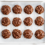 A top-down view of several no-bake chocolate oatmeal cookies placed neatly on a tray lined with parchment paper. The cookies are round, dark chocolate brown with visible oats, and slightly glossy from the peanut butter and cocoa mixture. They rest on a white marble countertop, cooled and ready to serve or store.