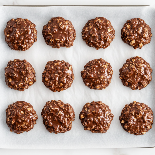 A top-down view of several no-bake chocolate oatmeal cookies placed neatly on a tray lined with parchment paper. The cookies are round, dark chocolate brown with visible oats, and slightly glossy from the peanut butter and cocoa mixture. They rest on a white marble countertop, cooled and ready to serve or store.