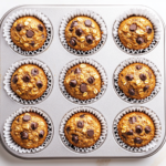 Top-down view of golden brown banana oat muffins with melted chocolate chips, arranged in white paper liners in a muffin pan. The muffins look soft and fluffy with a slightly crisp top. They are placed on a clean white marble countertop, and the scene is well-lit with a warm, homemade feel.