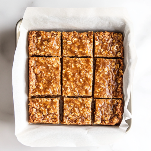Top-down image showing perfectly baked gingerbread oat bars sliced inside a parchment-lined 8-inch baking tin on a white marble countertop. The bars are golden brown with a shiny top and soft texture. Bits of candied ginger are visible, enhancing the homemade appearance. The clean setup highlights the rustic charm of this spiced dessert.