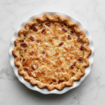 Top-down view of a freshly baked homemade apple pecan pie sitting in a white ceramic 9-inch pie dish on a white marble countertop. The pie has a golden flaky crust and is topped with a rich layer of glistening chopped pecans caramelized with brown sugar and margarine. The scene is clean, rustic, and inviting.