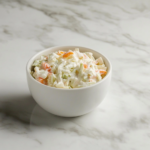 Top-down view of a bowl filled with creamy coleslaw made from chopped green cabbage, shredded carrots, and minced onion. The vegetables are coated in a rich dressing made from mayonnaise, vinegar, mustard, sugar, and pickles. The dish is served chilled in a white bowl over a clean marble background, capturing freshness and texture.