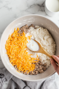 A large white mixing bowl on a white marble countertop, where a hand is pouring fresh milk from a small white measuring cup into the mixture of flour, cheese, sausage, and butter. The dough is starting to come together, slightly sticky but well combined. A wooden spoon is partially submerged in the mixture.