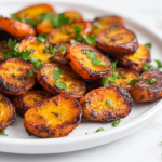 A Side view of perfectly roasted carrots served on a white ceramic plate. The carrots are golden brown and caramelized around the edges, showcasing their natural sweetness and roasted texture. Garnished with freshly chopped parsley, they rest on a clean white marble countertop, presenting a warm and rustic dish ideal for healthy meals.