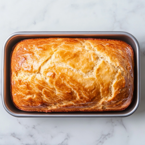Top-down view of a freshly baked banana bread loaf resting in a silver 9x5 inch metal pan placed on a clean white marble countertop. The loaf is golden brown with a cracked, slightly domed top, showcasing its soft and moist interior. The setup is simple and clean, highlighting the comforting, homemade feel of the banana bread.