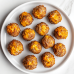 A plate filled with golden brown, freshly baked sausage cheese balls arranged on a white ceramic plate. The crispy, slightly cracked surface reveals melted cheddar cheese and flavorful sage sausage inside. The plate is placed on a white marble countertop with a minimal, clean background, making the delicious appetizer the main focus.