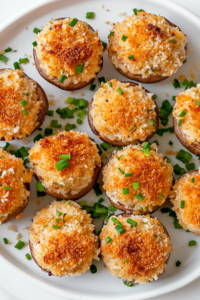 A white plate filled with golden brown stuffed mushrooms, generously packed with a creamy mixture of Italian sausage, cream cheese, garlic, and herbs. The tops are crisped with melted parmesan cheese and crunchy panko breadcrumbs. The plate sits on a white marble countertop, with fresh parsley and chopped chives sprinkled for garnish.