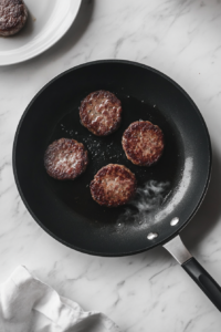 Several breakfast sausage patties are sizzling in a black non-stick skillet placed on a white marble countertop. A stainless steel spatula is flipping a patty, revealing a golden-brown seared surface. The patties have a slightly crisp outer texture while remaining juicy inside, releasing a delicious aroma as they cook.