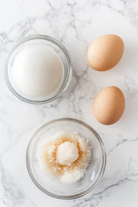 A top-down view showing three large eggs separated into two glass bowls. One bowl holds the yolks, mixed with granulated sugar and vanilla extract, while the other contains the egg whites. A whisk is placed next to the bowl of egg yolks, highlighting the preparation phase. The clean white marble countertop provides a minimalist background to emphasize the focus on separating and beating the eggs for the cake mixture.
