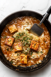A top-down view of ramen noodles, tofu, and sauce simmering together in a black skillet on a white marble countertop. The ingredients are blending together as they cook, with the sauce thickening and the flavors melding. The scene is clean and simple, showcasing the final cooking phase of the dish before serving.