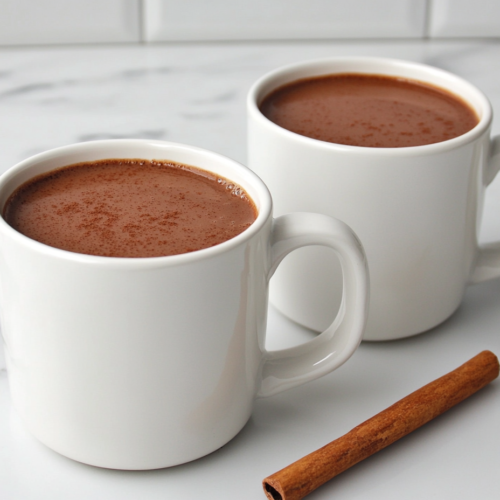 Top-down view of two white mugs filled with rich, creamy spiced hot chocolate, served on a white marble countertop. The hot chocolate has a deep brown color with a silky surface, garnished with a cinnamon stick resting on the mug rim. The setup is clean and elegant, capturing the cozy warmth of this homemade drink, ideal for winter evenings.