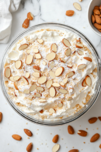 A top-down shot of fluffy, stiff-beaten egg whites being spread over the cake batter in the pans. Sliced almonds are gently sprinkled on top of the meringue. The glass mixing bowl of meringue and a spatula are visible, with the white marble countertop adding a clean and simple background. The scene focuses on the delicate assembly process for the Heaven Cake.