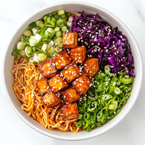 A top-down shot of a bowl of finished ramen, topped with crispy tofu, sautéed vegetables, sesame seeds, and chopped scallions, all served on a white marble countertop. The vibrant colors of the toppings contrast beautifully with the noodles and sauce, showcasing the dish in a simple yet inviting way. The clean setup draws attention to the completed, flavorful dish.