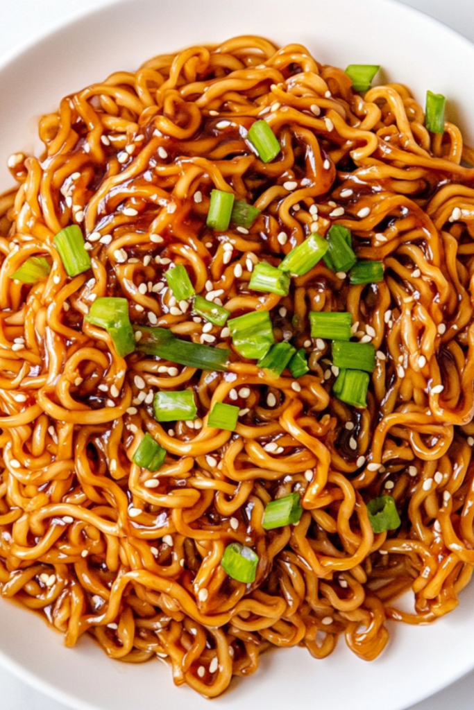 A top-down view of delicious teriyaki noodles served on a white ceramic plate, garnished with chopped green onions and sesame seeds. The noodles are perfectly coated in a glossy teriyaki sauce and look warm, comforting, and flavorful. This simple and quick meal is great for busy weeknights or casual lunches.