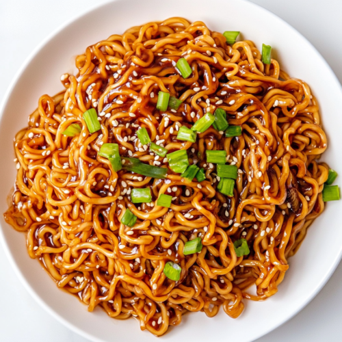 A top-down view of delicious teriyaki noodles served on a white ceramic plate, garnished with chopped green onions and sesame seeds. The noodles are perfectly coated in a glossy teriyaki sauce and look warm, comforting, and flavorful. This simple and quick meal is great for busy weeknights or casual lunches.