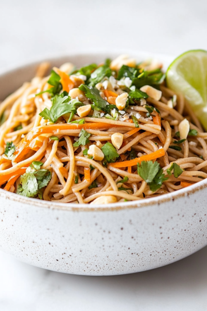 A top-down shot of a chilled soba noodle salad beautifully arranged in a white ceramic bowl, sitting on a clean white marble countertop. The noodles are mixed with finely grated carrots, fresh chopped cilantro, and sprinkled with coarsely chopped salted peanuts. A lime wedge is placed at the side, enhancing the fresh, zesty appearance of this vibrant and healthy salad.