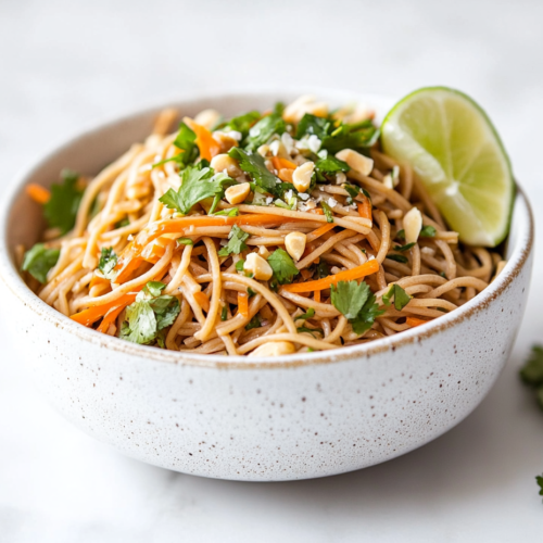 A top-down shot of a chilled soba noodle salad beautifully arranged in a white ceramic bowl, sitting on a clean white marble countertop. The noodles are mixed with finely grated carrots, fresh chopped cilantro, and sprinkled with coarsely chopped salted peanuts. A lime wedge is placed at the side, enhancing the fresh, zesty appearance of this vibrant and healthy salad.