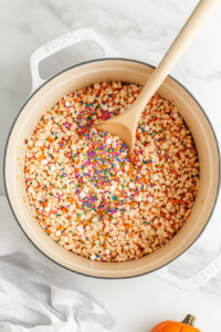 Top-down view of a large pot with Rice Krispies and marshmallow mixture, as Halloween-themed sprinkles are added and mixed gently with a wooden spoon, on a white marble countertop.