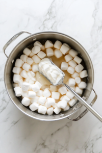 Top-down view of a large stainless steel pot with melted unsalted butter and marshmallows over low heat, gently stirred with a spatula, on a white marble countertop.