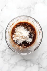 A top-down shot of a mixing bowl with a whisk, blending together vegetable broth, soy sauce, cornstarch, maple syrup, gochujang, vinegar, ground pepper, and sesame oil. The ingredients are well-combined, with a whisk in motion. The white marble countertop keeps the scene focused and uncluttered, ensuring the emphasis is on the sauce mixture.