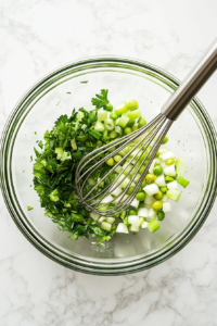 Freshly chopped vegetables and herbs are now added into the glass bowl filled with the seasoned sauce base. Ingredients include scallions, parsley, green olives, celery, and capers, creating a colorful contrast against the creamy orange background of the sauce. This step introduces texture and freshness to the remoulade, enhancing both the appearance and the flavor of the final result.