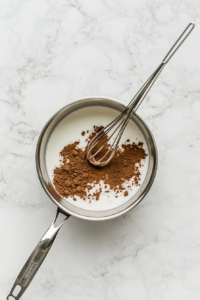 The image shows cocoa powder and sugar being added to the warm milk inside a medium silver saucepan placed on a white marble countertop. A stainless steel whisk stirs the ingredients, starting to blend the cocoa and sugar into the milk. The scene captures the transformation of plain milk into a rich chocolate base for the drink.