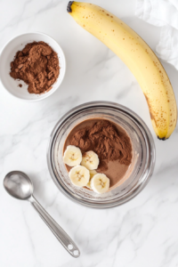 Cocoa powder is being added to a blender already containing almond milk, chopped banana, and peanut butter in this top-down view. The dark powder contrasts beautifully against the other ingredients in the blender. A small white bowl and measuring spoon are included in the scene, all resting on a clean white marble surface.