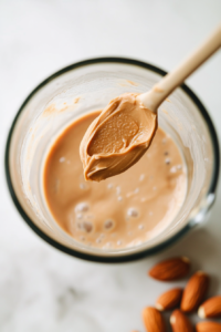 Top-down photo showing creamy peanut butter being added into a blender that already contains almond milk and chopped banana. A silver spoon holds the peanut butter just above the blender jar, ready to drop in. The countertop is white marble, creating a clean and consistent background that emphasizes each stage of preparation.