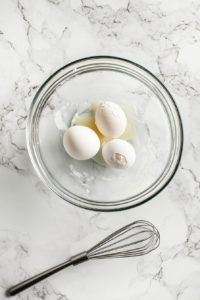 Top-down view of two cracked eggs and vanilla extract added into a sugar and butter mixture inside a clear glass bowl on a white marble surface. A metal whisk is partially submerged, ready to mix the ingredients. Eggshells and a teaspoon of vanilla rest nearby, capturing a crucial step in building flavor and richness for the banana bread batter.