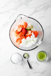 A blender filled with bright red frozen strawberries, white granulated sugar, vodka, and freshly squeezed lime juice. The ingredients are ready to be blended into a deliciously smooth and slushy frozen cocktail with a sweet and tangy flavor balance.