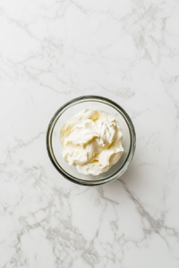 In this image, a clear glass bowl sits on a white marble countertop containing measured portions of mayonnaise and sour cream. These creamy base ingredients are the first step in creating the lemon garlic sauce. The clean background emphasizes the fresh ingredients and sets the tone for a simple and delicious dip preparation.