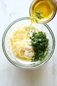 Finely chopped fresh parsley and chives, along with minced garlic and olive oil, are being added to the creamy mixture in the glass bowl. These ingredients enhance the flavor and color of the sauce. The setup shows each element going in clearly, demonstrating the layering of taste and freshness in the recipe.