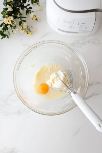 A large glass bowl on a white marble surface shows room-temperature eggs being added one at a time into a creamy mixture of butter and sweetener. A white electric hand mixer is used to blend the mixture thoroughly. Vanilla and pineapple extracts in small dishes are nearby, enhancing the flavor depth of the batter.