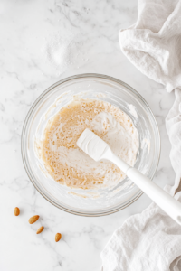 A hand pouring sour cream, almond extract, and vanilla extract into a bowl of whipped cream cheese. The mixture is being stirred together to create a luscious, aromatic blend that enhances the dessert’s flavor.