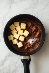 All the main wet and dry ingredients—white sugar, butter, milk, unsweetened cocoa powder, and a pinch of salt—are placed in a black saucepan, sitting on a white marble countertop. These ingredients form the chocolate base of the no-bake cookies. This mixture will be heated and stirred to create a smooth, rich syrup.