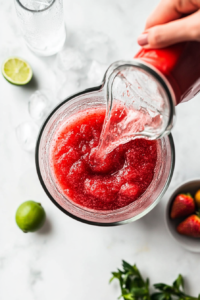 A hand pours a small amount of water or extra lime juice into the blender to adjust the thickness of the strawberry vodka slush. This step ensures the drink has the perfect smooth and icy consistency, making it easy to sip and enjoy.