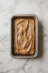 The banana bread batter has been poured into the greased 9x5 inch metal loaf pan and is shown ready for baking on a white marble countertop. The batter is smooth and evenly spread. This final preparation step is essential before placing the pan into a preheated oven at 350°F for baking.