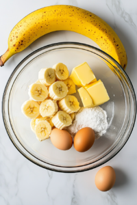 A top-down shot of a clear glass bowl filled with mashed bananas, granulated sugar, two large eggs, melted butter, and vanilla extract, sitting on a white marble kitchen counter. These wet ingredients are combined to create a rich, flavorful base for the banana bread. The setup is simple and neatly arranged.