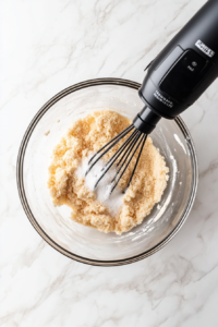 A close-up of a mixing bowl where softened cream cheese is being beaten with white sugar using an electric mixer. The mixture is turning smooth and creamy, ensuring a rich and velvety texture for the dessert base.