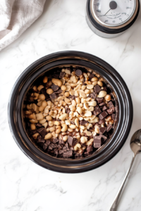 A black crock pot is covered with its lid and placed on a white marble countertop, slowly melting the layers of white and semisweet chocolate along with dry roasted peanuts inside. This slow cooking process on low heat helps the ingredients blend thoroughly, creating a smooth and creamy base for the clusters.