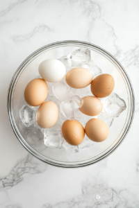 A clear glass bowl is filled with ice cubes and cold water, cooling the just-boiled eggs quickly. This shocking method helps stop the cooking process and makes peeling much easier. Set on a bright white marble countertop, the image reflects a clean, simple kitchen setup during this vital cooling stage.