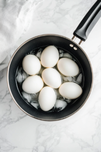 Freshly boiled eggs are cooling in a clear glass bowl filled with ice cubes and water, placed on a white marble surface. This ice bath helps stop the cooking process and makes peeling the eggs much easier. It ensures that the egg whites remain tender while the yolks stay creamy, not overcooked or rubbery.