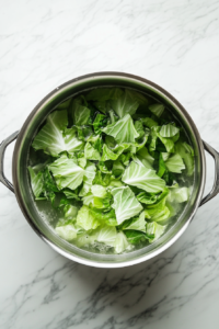 Top-down image of a large pot on a white marble countertop where pale green cabbage leaves are being blanched in boiling water. Tongs rest nearby, indicating the leaves are being softened for wrapping. This crucial step ensures the cabbage becomes pliable enough to roll tightly around the meat and rice filling in the later steps.