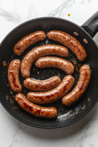 Top-down shot of a black skillet on a white marble surface with 1 pound of bulk sausage being cooked and crumbled over medium heat. The sausage is browned and sizzling, with a wooden spoon beside it. This key step adds hearty flavor to the quiche mixture, and excess fat is drained after cooking.