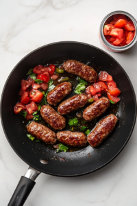 Top-down view of a black skillet filled with browned ground breakfast sausage combined with diced tomatoes and green chiles. The skillet is on a white marble countertop. A spatula rests beside the skillet. The ingredients are sizzling together, creating a flavorful base for the casserole with steam slightly rising from the mixture.