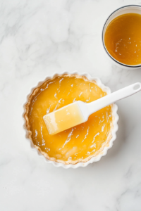 The tart is baking in a white pan on a white marble countertop. The apple slices are softening while the custard sets to a creamy consistency in the oven at 350°F. The surface is gradually becoming golden and firm. This stage is key for melding flavors and textures together to create a balanced tart. The image captures the mid-bake moment before final finishing touches are added.