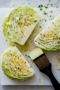 Cabbage steaks are being brushed generously with light olive oil and freshly minced garlic using a black-handled basting brush. The garlic and oil mixture adds rich flavor and prevents sticking on the grill. Placed on a white cutting board over a white marble counter, this scene captures the crucial flavor-enhancing preparation before grilling the cabbage steaks to perfection.