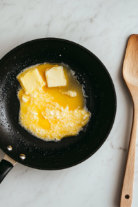 A top-down shot of butter and sesame oil melting together in a black skillet placed on a white marble cooktop. The butter is bubbling slightly, beginning to combine with the sesame oil, creating a flavorful base for stir-frying. The wooden spatula lies next to the skillet. This step adds richness and nutty depth to the noodle dish.